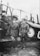 Two men in military uniforms stand in front of a vintage biplane. They are wearing caps and knee-high boots, and both display badges on their jackets. The biplane behind them has a roundel emblem on its side.