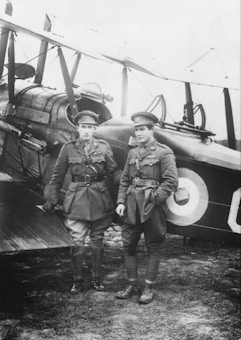 Two men in military uniforms stand in front of a vintage biplane. They are wearing caps and knee-high boots, and both display badges on their jackets. The biplane behind them has a roundel emblem on its side.