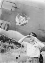 A smiling man in a military uniform stands saluting in front of an airplane. The airplane has a cartoon character painted on its side near the man's head.