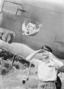 A smiling man in a military uniform stands saluting in front of an airplane. The airplane has a cartoon character painted on its side near the man's head.