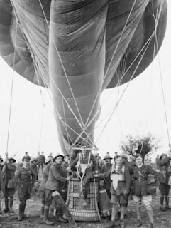 A group of military personnel dressed in early 20th-century uniforms gather around a large tethered balloon. The balloon is inflated with ropes securing it. One man sits on the edge of the balloon's basket.