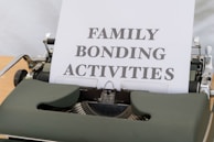 A typewriter with vintage design holding a sheet of paper that has the words 'FAMILY BONDING ACTIVITIES' typed in bold, black font. The typewriter is a dark green color, and the sheet of paper is positioned upright, indicating its recent use.