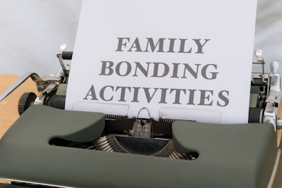 A typewriter with vintage design holding a sheet of paper that has the words 'FAMILY BONDING ACTIVITIES' typed in bold, black font. The typewriter is a dark green color, and the sheet of paper is positioned upright, indicating its recent use.