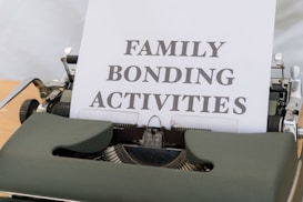 A typewriter with vintage design holding a sheet of paper that has the words 'FAMILY BONDING ACTIVITIES' typed in bold, black font. The typewriter is a dark green color, and the sheet of paper is positioned upright, indicating its recent use.