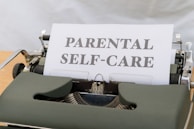 A close-up image of a vintage typewriter with a single sheet of paper inserted, displaying the words 'PARENTAL SELF-CARE' in bold letters. The typewriter has a classic design with visible keys and a dark green body.