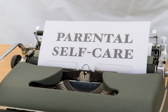 A close-up image of a vintage typewriter with a single sheet of paper inserted, displaying the words 'PARENTAL SELF-CARE' in bold letters. The typewriter has a classic design with visible keys and a dark green body.