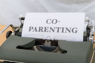A close-up view of a vintage typewriter displaying a piece of paper with the word 'CO-PARENTING' typed in bold letters. The typewriter has a dark green cover and is placed on a light wooden desk with a neutral background.