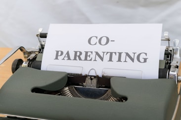 A close-up view of a vintage typewriter displaying a piece of paper with the word 'CO-PARENTING' typed in bold letters. The typewriter has a dark green cover and is placed on a light wooden desk with a neutral background.
