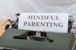 A vintage typewriter with a green body holds a sheet of paper displaying the words 'MINDFUL PARENTING' in bold, uppercase letters. The typewriter is placed on a light wooden surface, creating a classic and professional feel.