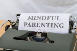 A vintage typewriter with a green body holds a sheet of paper displaying the words 'MINDFUL PARENTING' in bold, uppercase letters. The typewriter is placed on a light wooden surface, creating a classic and professional feel.