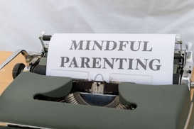 A vintage typewriter with a green body holds a sheet of paper displaying the words 'MINDFUL PARENTING' in bold, uppercase letters. The typewriter is placed on a light wooden surface, creating a classic and professional feel.