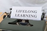 A typewriter with a sheet of paper displaying the words 'LIFELONG LEARNING' in bold letters. The typewriter appears vintage with a green cover, showcasing mechanical components around the paper.