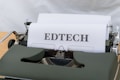A vintage typewriter with a sheet of paper that has the word 'EDTECH' typed on it. The typewriter casing is dark green and the setting appears to be on a light wooden surface.