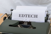 A vintage typewriter with a sheet of paper that has the word 'EDTECH' typed on it. The typewriter casing is dark green and the setting appears to be on a light wooden surface.