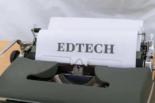 A vintage typewriter with a sheet of paper that has the word 'EDTECH' typed on it. The typewriter casing is dark green and the setting appears to be on a light wooden surface.