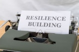 A vintage typewriter holds a sheet of paper with the words 'RESILIENCE BUILDING' typed in bold font. The typewriter is an older model with dark green keys and a metallic structure. The paper is crisp and white, contrasting with the machine's darker features.