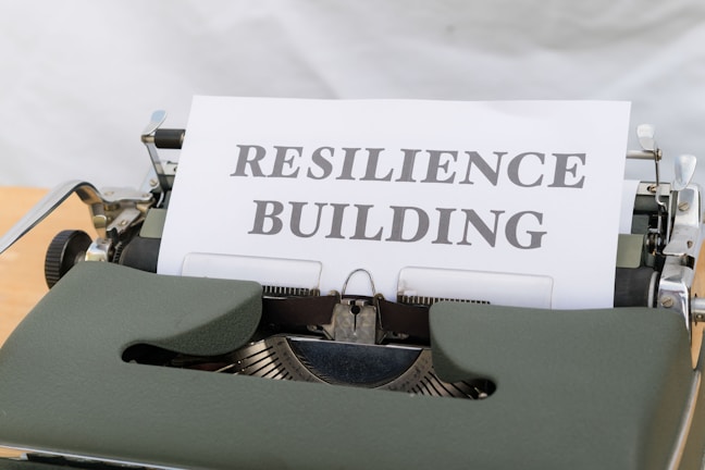 A vintage typewriter holds a sheet of paper with the words 'RESILIENCE BUILDING' typed in bold font. The typewriter is an older model with dark green keys and a metallic structure. The paper is crisp and white, contrasting with the machine's darker features.