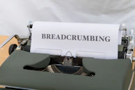 A vintage typewriter with a green casing is displaying a sheet of paper that has the word 'BREADCRUMBING' typed in bold uppercase letters. The typewriter is placed on a wooden surface, and the background appears to be a soft, neutral color.