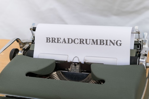 A vintage typewriter with a green casing is displaying a sheet of paper that has the word 'BREADCRUMBING' typed in bold uppercase letters. The typewriter is placed on a wooden surface, and the background appears to be a soft, neutral color.
