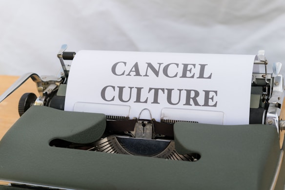 A vintage typewriter with paper showing the words 'CANCEL CULTURE' in bold, black letters. The typewriter has a dark green body, and the background is a soft white cloth.