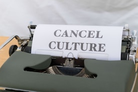 A vintage typewriter with paper showing the words 'CANCEL CULTURE' in bold, black letters. The typewriter has a dark green body, and the background is a soft white cloth.