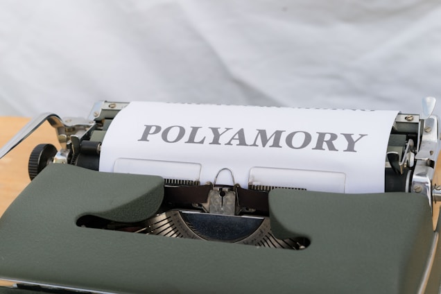 A close-up view of a typewriter with a sheet of paper displaying the word 'POLYAMORY' in bold letters. The typewriter has a dark green cover and is set against a light background.