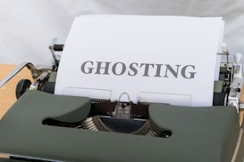 A vintage typewriter with a sheet of paper inserted. The word 'GHOSTING' is typed in bold capital letters on the paper. The typewriter has a dark green body and is set against a neutral backdrop, creating a nostalgic and classic look.