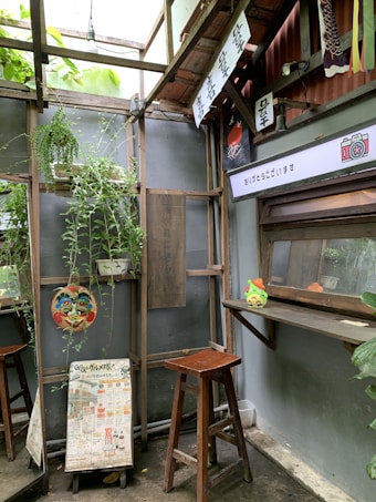 A cozy corner decorated with various elements, including wooden stools and shelves. Lush green plants hang and grow against a backdrop of a wooden structure. Colorful signs with Japanese characters adorn the area, along with artistic elements like a mask. A printed menu or pamphlet is displayed leaning against the wall.