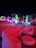 A vibrant outdoor scene featuring modern, colorful light sculptures and illuminated furniture at night. The environment includes creatively shaped light installations, glowing orbs, and contemporary seating arrangements, all bathed in vivid hues of red, green, blue, and purple. Flags and plants can be seen in the background, enhancing the atmosphere.