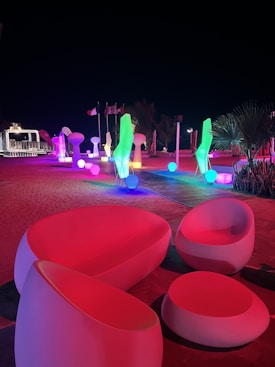 A vibrant outdoor scene featuring modern, colorful light sculptures and illuminated furniture at night. The environment includes creatively shaped light installations, glowing orbs, and contemporary seating arrangements, all bathed in vivid hues of red, green, blue, and purple. Flags and plants can be seen in the background, enhancing the atmosphere.