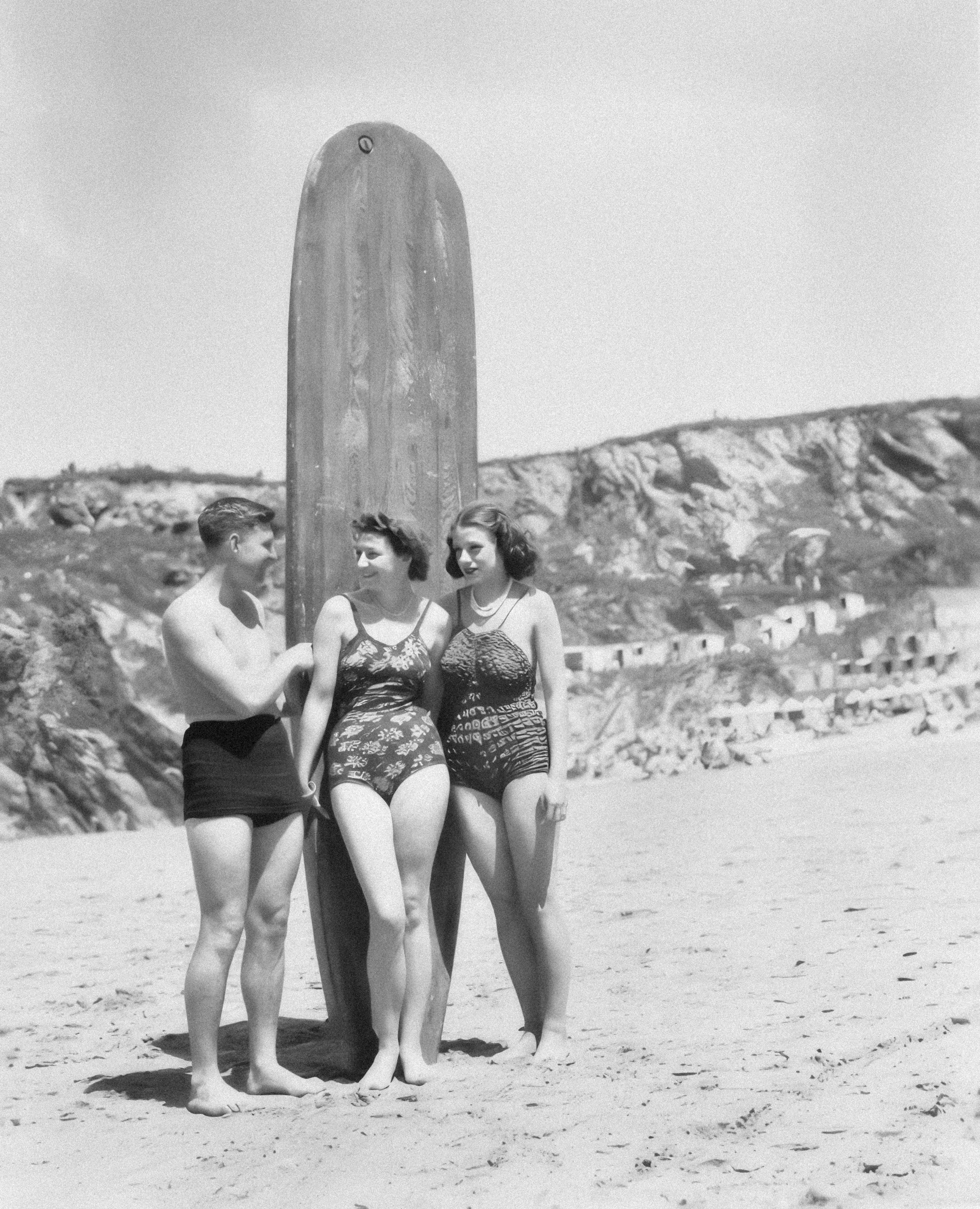 two women and a man standing on a beach with a surfboard