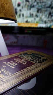 A richly decorated, embossed book with ornate golden patterns and Arabic script, lying on a surface. In the background, an illuminated lamp with a floral design casts a soft light, and a blurred image on a screen shows a crowd or gathering.