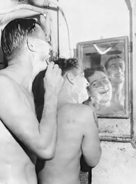 Two men are shaving in front of a small mirror. One is using a straight razor while the other gazes into the mirror, both with faces covered in shaving foam. The setting appears to be a rustic or vintage bathroom.