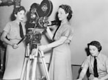 Three women are engaged in operating film equipment. Two of them are standing next to a vintage movie camera, while the third is seated and appears to be monitoring or taking notes. They are wearing uniforms typical of a mid-20th century setting.