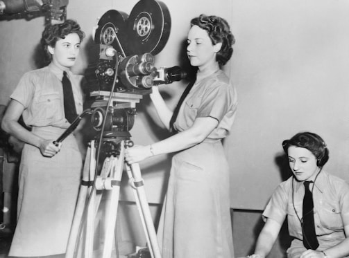 Three women are engaged in operating film equipment. Two of them are standing next to a vintage movie camera, while the third is seated and appears to be monitoring or taking notes. They are wearing uniforms typical of a mid-20th century setting.