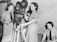 Three women are engaged in operating film equipment. Two of them are standing next to a vintage movie camera, while the third is seated and appears to be monitoring or taking notes. They are wearing uniforms typical of a mid-20th century setting.