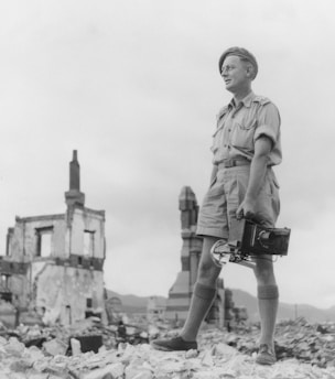 A person in vintage military attire holding a large camera stands amidst the rubble of a building in a desolate landscape. Remnants of structures are visible in the background, creating a stark contrast with the overcast sky.