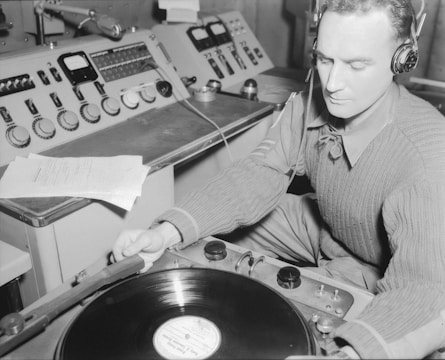A person wearing headphones operates a record player in a vintage sound control room. The setting includes an array of dials and meters on a large console with scattered papers. The individual is focused on the task, suggesting a technical or broadcasting environment.