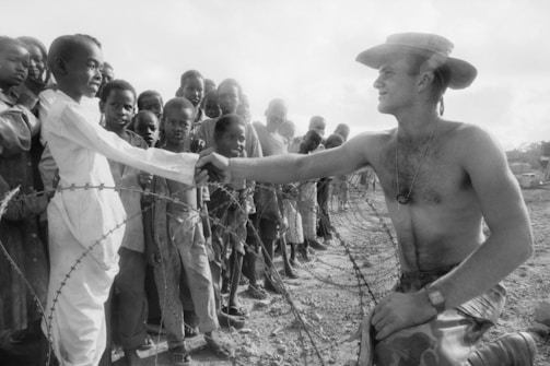 A shirtless man wearing a hat kneels next to a barbed wire fence, shaking hands with a young boy who is accompanied by a group of children standing behind him. The scene conveys a sense of connection despite the barrier. The setting appears to be outdoors under natural lighting.