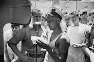 A group of sailors gather around a central figure reading from a piece of paper, who is dressed in a sleeveless shirt and sailor cap. The scene appears to be taking place on a ship, with a large bulkhead visible in the background. The group seems attentive and focused, suggesting an important announcement or event. Tattoos are visible on the arm of the central figure.