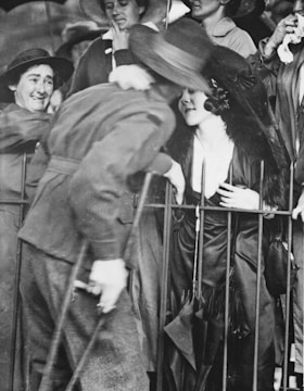 A black-and-white photograph capturing a lively group of people behind a metal barricade. The central figure is a woman wearing an elegant outfit with a large hat and fur stole, leaning towards a man holding onto the railing with crutches. The scene is crowded, with faces expressing a mix of curiosity and engagement.