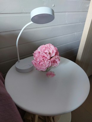 A small white table with a modern desk lamp and a bouquet of pink flowers in a glass container. One flower petal is lying on the table. The background consists of light wooden panels, contributing to a minimalist and serene ambiance.