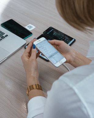 A person holding a smartphone displaying a settings menu, with two additional phones, a laptop, and a QR code placed on a light wooden table. The individual is wearing a gold watch and a white shirt.