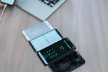 Several smartphones are placed in a row on a wooden surface, with a laptop partially visible in the background. The phones display various apps and screens, indicating usage and functionality. A person's hand can be seen holding another smartphone in the top left corner.