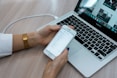 A person holds a smartphone displaying a checklist while sitting at a desk with a laptop open. The laptop screen shows multiple applications, with a focus on productivity tools. The desk surface is light wood, and the person is wearing a white shirt with a gold watch.