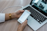 A person holds a smartphone displaying a checklist while sitting at a desk with a laptop open. The laptop screen shows multiple applications, with a focus on productivity tools. The desk surface is light wood, and the person is wearing a white shirt with a gold watch.