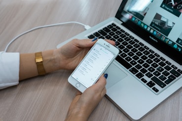 A person holds a smartphone displaying a checklist while sitting at a desk with a laptop open. The laptop screen shows multiple applications, with a focus on productivity tools. The desk surface is light wood, and the person is wearing a white shirt with a gold watch.