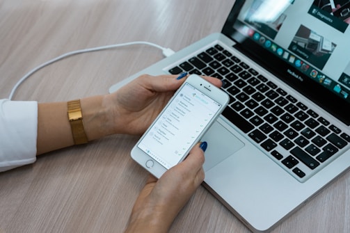 A person holds a smartphone displaying a checklist while sitting at a desk with a laptop open. The laptop screen shows multiple applications, with a focus on productivity tools. The desk surface is light wood, and the person is wearing a white shirt with a gold watch.