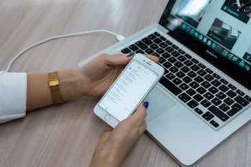 A person holds a smartphone displaying a checklist while sitting at a desk with a laptop open. The laptop screen shows multiple applications, with a focus on productivity tools. The desk surface is light wood, and the person is wearing a white shirt with a gold watch.