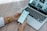 A person is holding a smartphone displaying a to-do list or task management app near an open laptop. The phone is connected to the laptop with a cable. The laptop is on a wooden desk showing web pages on the screen. The person is wearing a white sleeve and a gold watch.